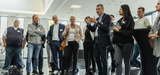 Gruppenfoto mit vielen Leuten, die in einem Raum zusammenstehen und diskutieren.