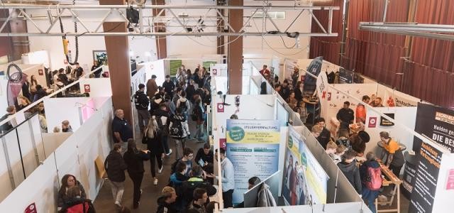 Blick von oben in eine Halle mit mehreren Messeständen und Besucherströmen.