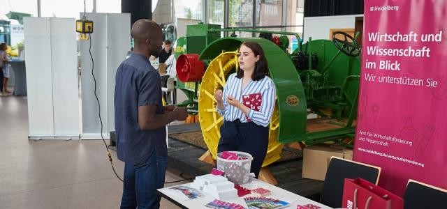 Eine Frau unterhält sich mit einem Mann. Sie steht an einem Stand mit Broschüren und Giveaways.