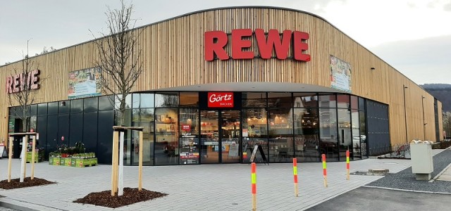 Ein Gebäude mit Holzfassade. Es hängen die Buchstaben REWE über dem Eingang.