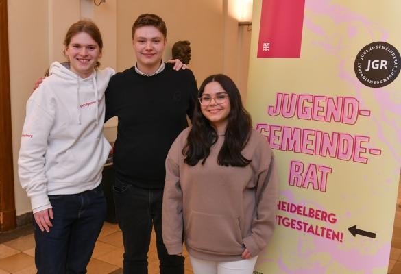 Der neue Vorstand des Heidelberger Jugendgemeinderats (drei Personen) steht vor einem Werbeplakat