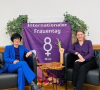 Bürgermeisterin Stefanie Jansen auf der Couch sitzend neben der Gleichstellungsbeauftragten der Stadt Heidelberg, zwischen ihnen ein Banner für den internationalen Frauentag 