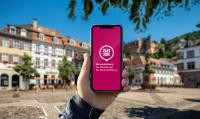 Das Rathaus für die Hosentasche: Mit der „MeinHeidelberg“-App haben Bürgerinnen und Bürger ebenso wie Besucherinnen und Besucher eine geballte Ladung an Services und Informationen immer dabei. (Foto: Stadt Heidelberg) Hochgehaltenes Handy mit der App vor dem Marktplatz