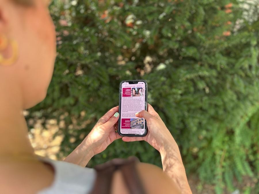 Die neuen Stadtteil-Rallyes in der MeinHeidelbergApp führen an Orte, die für junge Menschen interessant sind. (Foto: Stadt Heidelberg) Blick über die Schulter einer jungen Frau, sie bedient ein Smartphone mit geöffneter MeinHeidelbergApp, im Hintergrund ist verschwommen ein Busch zu sehen.