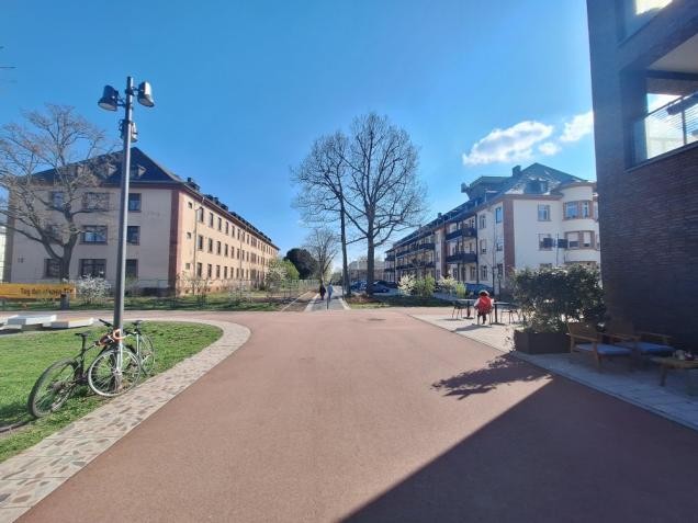 Das verkaufte Gebäude (links) liegt zentral in der Nähe des Paradeplatzes und am Fußweg der Adelheid-Steinmann-Straße. Es wird künftig die Heimat des gemeinschaftlichen Wohnprojektes paraSol sein. (Foto: Entwicklungsgesellschaft Campbell Barracks mbH) Zu sehen ist ein breiter Weg der nach links abbiegt. Weiter geradeaus liegen zu beiden Seiten 2 große Gebäude.