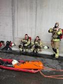 Zwei Feuerwehrleute sitzen nach einer Übung auf dem Boden und trinken. Sie lachen in die Kamera. Ein weiterer Feuerwehrmann zieht seine Uniform an und macht sich für die Übung bereit.