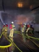 Vier Feuerwehrangehörige während der Übung. Ein Trupp kühlt mit Wasser die Tunnelwände, ein anderer Trupp löscht ein brennendes Auto ab. Ein fünfter Feuerwehrmann beobachtet die beiden Trupps.