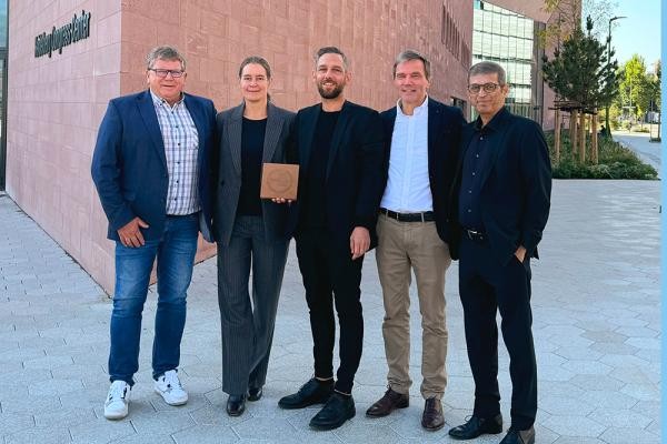 Fünf Personen stehen vor einem Gebäude. Eine hält einen Preis in der Hand.