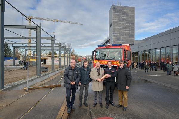 Fünf Personen, darunter ein Feuerwehrmann, stehen auf einem Baustellengelände und halten einen zylindrischen Behälter aus Kupfer.