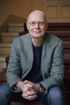Jens Siegert A man in his 60s sits on a staircase with his hands folded. He looks at us confidently .