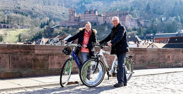 Eine Frau und ein Mann stehen mit zwei Fahrrädern auf der Alten Brücke. Im Hintergrund ist das Schloss und die Sonne scheint. (Foto: Stadt heidelberg/ Pellner)