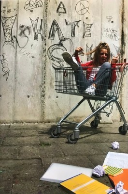 Ein Foto von Hannah in einem Einkaufswagen vor einer besprayten Wand. Im Vordergrund liegen Hefte, ein Buch und zerknülltes Papier.