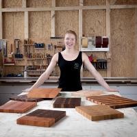Schreinermeisterin Maxime Krämer zeigt stolz die individuell gestaltbaren hochwertigen Holzschneidebretter, die Kunden bei ihr beauftragen können. (Bild: Krämer) Maxime Krämer steht am Tisch vor ihren selbstgeschreinerten Schneidebrettern.