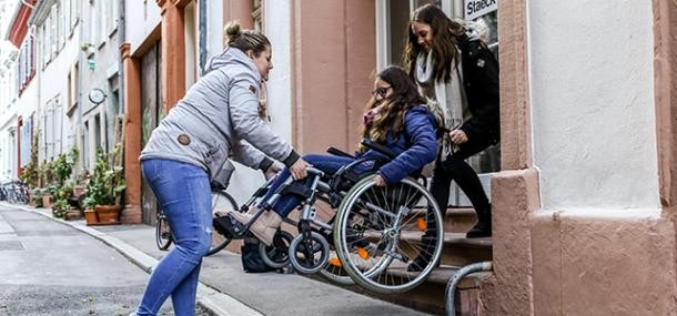 Zwei Frauen tragen eine Frau im Rollstuhl eine Treppe hinunter