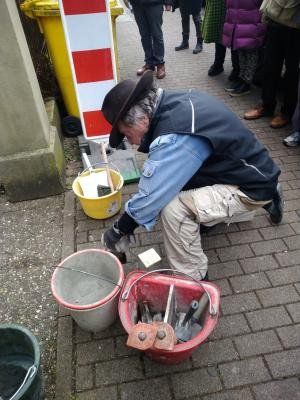 Gunter Demnig in Arbeitskleidung und mit Hut fügt den messingfarbenen Stolperstein in den Gehwegbelag ein