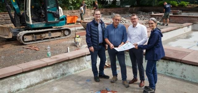 Bürgermeister Raoul Schmidt-Lamontain steht mit Andreas Lippke, Joachim Becker und Karen Engelhardt im Brunnenbecken, neben mehreren rot-blauen Fisch-Mosaiken..