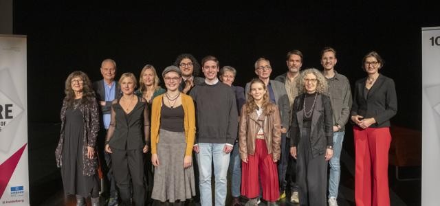 15 Menschen stehen eingerahmt von Roll-ups auf einer Bühne.