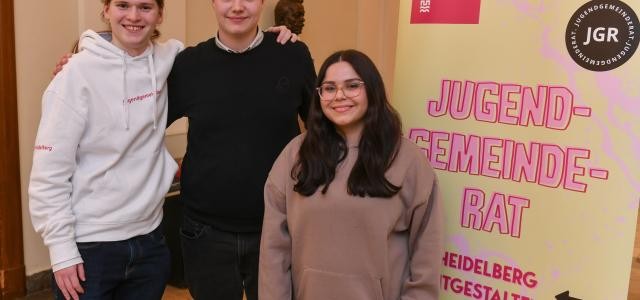 Der neue Vorstand des Heidelberger Jugendgemeinderats (drei Personen) steht vor einem Werbeplakat
