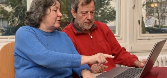 Zwei ältere Menschen (eine Frau in blauem Pulli und ein mann in rotem Pulli) sitzen mit einem Laptop an einem Tisch. Die Frau zeigt mit ihrem Finger auf den Bildschirm, beide schauen in die Richtung des Bildschirmes.