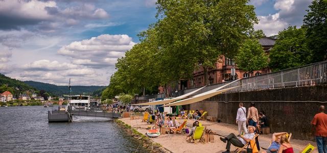 Auf einem aufgeschütteten Strand am Flussufer liegen eini´ge Personen auf Liege, andere flanieren vorbei.