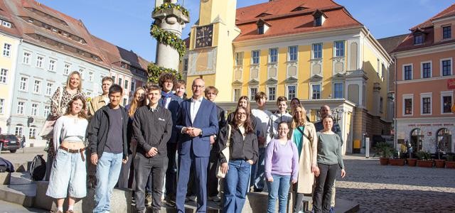 Eine Gruppe junger Menschen mit einem mittelalten Herrn in Anzug stehen vor einem Brunnen in malerischer Altstadtkulisse.