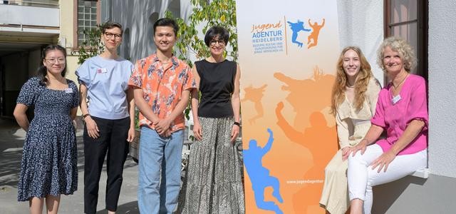 Sechs Menschen stehen vor einem Gebäude neben einem orangefarbenen Roll-up in der Sonne