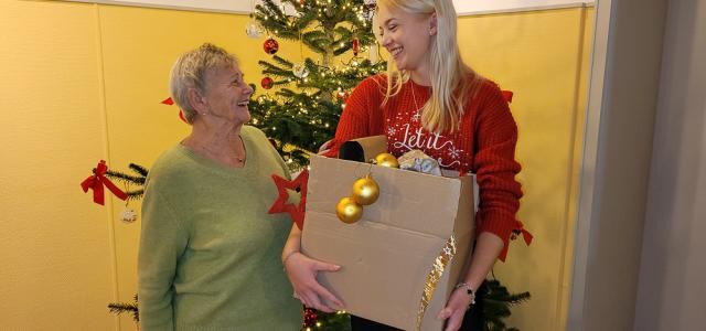 Eine ältere und eine junge Frau stehen vor einem Weihnachtsbaum. Die junge Frau hält einen Karton voller Weihnachtsschmuck.