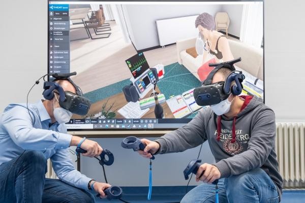 Zwei Männer haben VR-Brillen auf und Controller in der Hand. Hinter ihnen ist ein Bildschirm zu sehen.