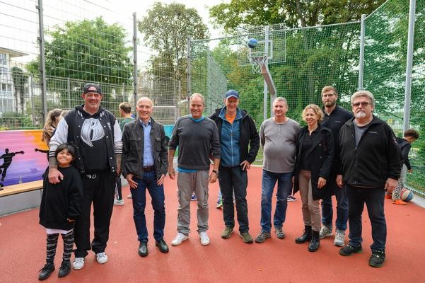 Freuen sich über die Eröffnung des neuen Streetballplatzes in Bergheim: Michel Vogt mit Tochter, Volker Schwarz (Leiter Abteilung Grünanlagen beim Landschafts- und Forstamt), Oberbürgermeister Eckart Würzner, Dr. Ernst Baader (Amtsleiter Landschafts- und Forstamt), Alfred Szpalecki (Regiebetrieb Gartenbau beim Landschafts- und Forstamt), Angelika Kern (Abteilung Grünanlagen, Fachbereichsleiterin Spiel- und Freizeitanlagen beim Landschafts- und Forstamt, Frederick Wetzel (Garten- und Landschaftsbau), Uli Hartmann (Ballfangnetz) (v.l.). (Foto: Stadt Heidelberg) Eine Gruppe von Menschen steht auf dem neuen Streetballplatz. Im Hintergrund ist der Korb mit einem Ball zu sehen.
