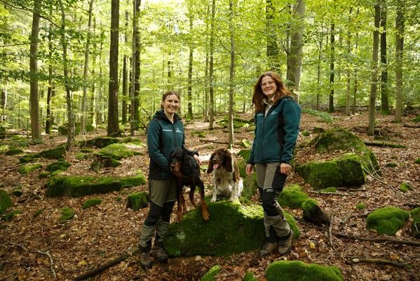 Försterin Charlotte Schulz (l.) und Wildtierbeauftragte Luisa Krauß mit ihren Kadaverspürhunden Ava und Lucie. (Foto: Stadt Heidelberg) Zwei Frauen stehen in Outdoorkleidung im Wald mit ihren Hunden.