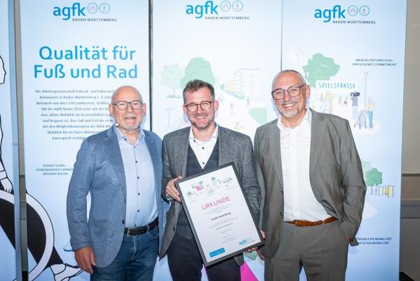 Mobilitätsbürgermeister Raoul Schmidt-Lamontain (vorne, 2. v.l) nahm für die Stadt Heidelberg am 25. Oktober 2024 die Auszeichnung entgegen. Zudem wurde er in Tübingen auch in den Vorstand der AGFK gewählt. (Foto: AGFK BW) Drei Männer stehen mit einer Urkunde vor einem großen Roll-up.