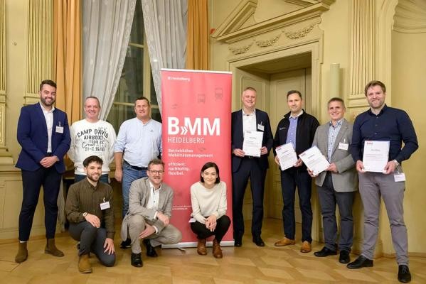 Bei der Abschlussveranstaltung zum Betrieblichen Mobilitätsmanagement (BMM) am 11. November 2024 überreichte Klima- und Mobilitätsbürgermeister Raoul Schmidt-Lamontain (vorne, 2. von links) Unternehmensvertretern der vier diesjährigen Teilnehmer (rechts) ihre Teilnahmezertifikate. (Foto: Philipp Rothe) Gruppe von Menschen vor und neben dem roten BMM-Aufsteller