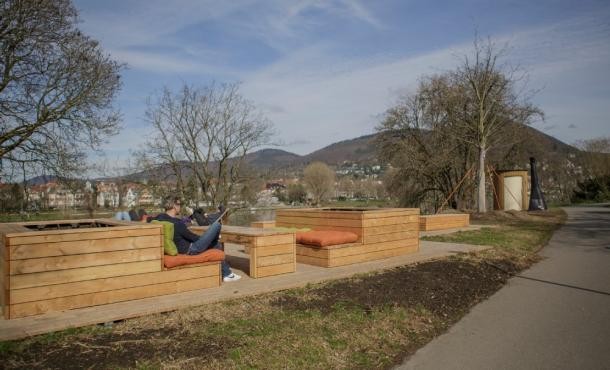 Das Iqbal-Ufer in Bergheim wurde 2024 durch das Mittendrinnenstadt-Programm aufgewertet – neben einer öffentlichen Toilette wurde ein Poesie-Automat aufgestellt und ein Strom- und Wasseranschluss für Veranstaltungen gelegt. Betrieben wird der „Neckarort“ durch den Verein Neckarorte e.V. (Foto: Buck) Holzbänke auf einer grünen Wiese.