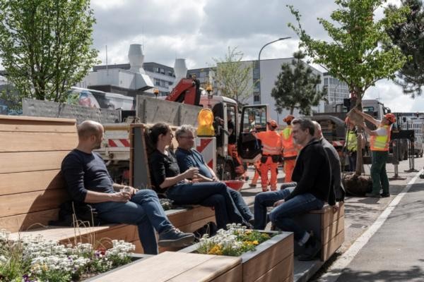 Die Emil-Maier-Oase beim Kultur- und Kreativwirtschaftszentrum Dezernat 16 lädt zum Verweilen und Erholen ein. Im Frühjahr 2024 wurden dort verschiedene Pflanzen in Kübeln aufgestellt – ein Parklet ermöglicht beispielsweise entspannte Mittagspausen und Feierabende. (Foto: Buck) Eine Gruppe Menschen sitzen auf Holzbänken und unterhalten sich. Im Hintergrund sieht man eine Baustelle.