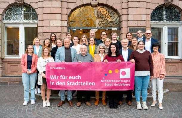 Aktiv für Kinderinteressen in den Heidelberger Stadtteilen: Die Kinderbeauftragten mit Oberbürgermeister Eckart Würzner (Reihe vorne 3.v.l.) und Bürgermeisterin Stefanie Jansen (Reihe vorne 1.v.r.). (Foto: Bäcker) Guppenfoto einer Guppe Menschen, die einen Banner vor sich halten.