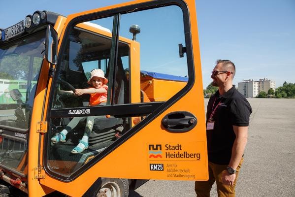 In die Kabine einer Kehrmaschine zu klettern, ist für die Kleinen gar nicht so einfach. Dafür ist es am Lenkrad umso schöner. (Foto: Stadt Heidelberg) Ein kleine Mädchen sitzt am Lenkrad eines orangen Lastwagens, neben ihr ein Mann mit Sonnenbrille.