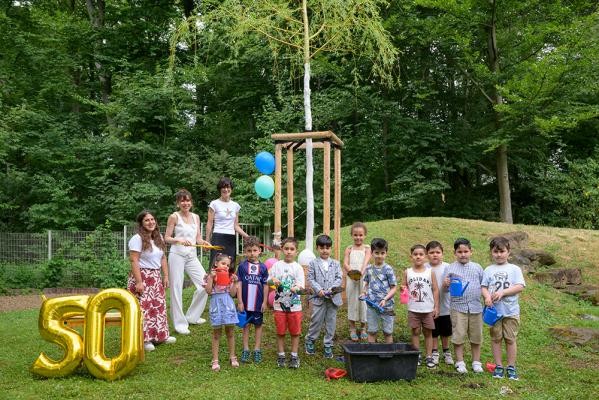 Gemeinsam wachsen: Die frisch gepflanzte Trauerweide im Garten der Kita Buchwaldweg soll als Geburtstagsgeschenk dauerhaft an das 50. Jubiläum erinnern. Beim Sommerfest wurde der Baum von den Kita-Kindern gemeinsam mit Bürgermeisterin Stefanie Jansen (3. von links) und den Kita-Leiterinnen Ayla Arbaz (2. von links) und Songül Uluca (links) symbolisch mit Erde bedeckt und angegossen. (Foto: Rothe) Drei Frauen und einige Kinder stehen um einen jungen Baum; an der Seite sind Ballone in der Form einer 50 zu sehen.