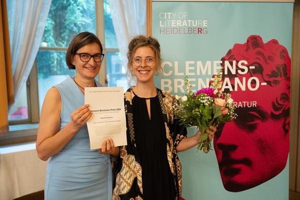 Erhielt den Clemens-Brentano-Preis der Stadt Heidelberg 2025: Noemi Somalvico. (Foto: Gös) Zwei Frauen halten strahlend eine Urkunde und einen Blumenstrauß in den Händen, im Hintergrund ist das türkise Plakat des Brentano-Preis zu sehen.