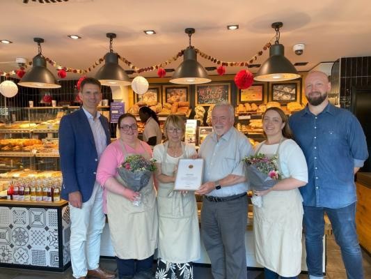 Marc Massoth (von links), Leiter des Amtes für Wirtschaftsförderung und Wissenschaft, gratulierte dem Familienbetrieb mit Geschäftsführerin Sabine Frisch, den beiden Senior-Chefs Karin Frisch und Wolfgang Frisch, Geschäftsführerin Sandra Frisch und Alexander Frisch. (Foto: Stadt Heidelberg) Marc Massoth, Sabine Frisch, Karin Frisch, Wolfgang Frisch, Sandra Frisch und Alexander Frisch stehen mit zwei Blumensträußen und der Urkunde vor dem Verkaufstresen im Café Frisch.