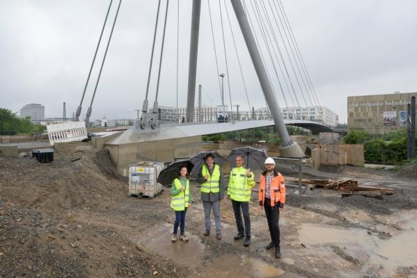 500 Tonnen Stahl werden von 24 Drahtseilen und dem Pylon getragen: Die Gneisenaubrücke ist kurz vor der Fertigstellung und bietet ab nächstem Jahr für alle Radfahrende, Fußgängerinnen und Fußgänger eine komfortable Querung der Gleise zwischen Bergheim und Bahnstadt (Hintergrund). Bei der Besichtigung vor Ort (von links): Kerstin Maixner (Geschäftsstelle Bahnstadt), Erster Bürgermeister Jürgen Odszuck, Klaus-Peter Hofbauer (Leiter Tiefbauamt) und Vinzenz Borchert (Projektleiter im Tiefbauamt). Jürgen Odszuck, Andrea Maixner, Klaus-Peter Hofbauern und Vinzenz Borchert stehen im Baustellenbereich der Gneisenaubrücke, hinter ihnen sieht man bereits einen großen Teil der Brücke.