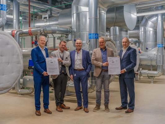 Freude über zwei Förderbescheide in Höhe von insgesamt 86 Millionen Euro für die Projekte Flusswärmepumpe, Abwasserwärmepumpe und Ausbau des Fernwärmenetzes (von links): Falk Günther (Geschäftsführer Stadtwerke Heidelberg Netze), Raoul Schmidt-Lamontain (Bürgermeister für Klimaschutz, Umwelt und Mobilität), Prof. Dr. Rudolf Irmscher (Geschäftsführer Stadtwerke Heidelberg), Oberbürgermeister Eckart Würzner und Michael Teigeler (Geschäftsführer Stadtwerke Heidelberg Energie) in der innovativen Kraft-Wärme-Kopplungsanlage, die bereits heute zur klimaneutralen Fernwärmeversorgung beiträgt. (Foto: Tobias Dittmer) 5 Personen stehen in einem industriell aussehenden Raum mit silbernen Rohren und präsentieren 2 Plakate.