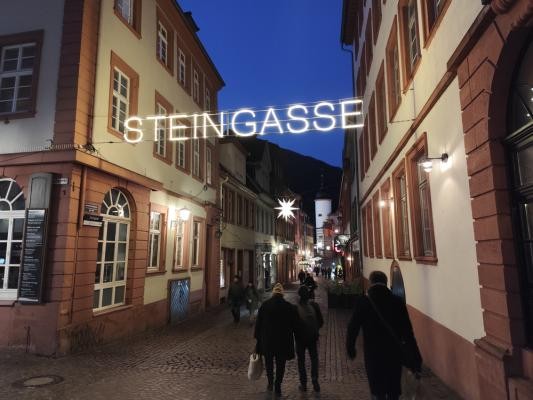 Fünf Seitengassen wurden mit beleuchtenden Schriftzügen aufgewertet und laden zum Einkaufen ein – darunter die Steingasse (Foto: Stadt Heidelberg) zu sehen ist eine leuchtender Schriftzug mit der Aufschrift STEINGASSE der über der benannten Straße hängt. Es ist dunkel und Menschen laufen durch die Straßße.