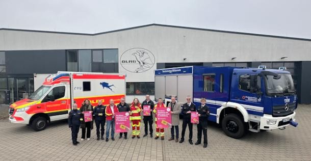 Die Ehrenamtskarte kommt (Foto: Stadt Heidelberg) Gruppenfoto von Angehörigen von Feuerwehr, DLRG und THW mit OB Würzner vor Einsatzfahrzeugen. Die Teilnehmer halten Infoplakate zu Ehrenamtskarte in die Kamera.
