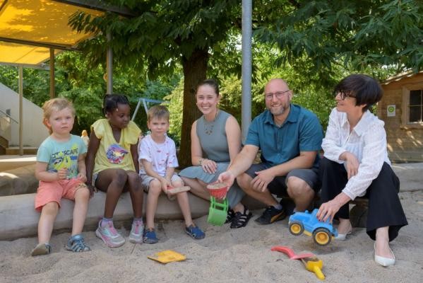 Die Kolumbianerin Andrea Gamboa (Mitte) ergänzt seit Jahresbeginn das Team in der Kirchheimer Kita Im Hüttenbühl. Bürgermeisterin Stefanie Jansen (r.) besuchte sie auf ihrer Sommertour. Auch der stellvertretende Kita-Leiter Sven Weiß (2.v.r.) und die Kinder der Kita freuen sich über viele neue Impulse, die sie aus ihrem Heimatland mitbringt. (Foto: Philipp Rothe) Drei Kinder und drei Erwachsene sitzen gemeinsam im Außenbereich einer Kindertagesstätte.