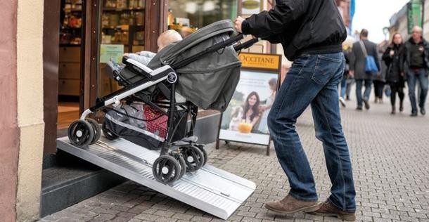 Ein Mann schiebt ein Kind im Kinderwagen  über eine Rampe in ein Ladengeschäft