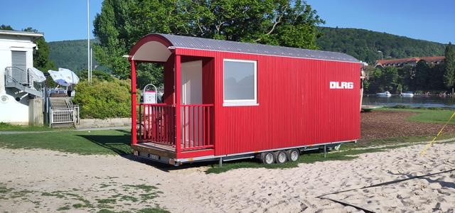 Ein hölzerner, leuchtend roter Wagen mit Aufschrift DLRG steht auf einer Wiese neben einem Beachvolleyballfeld.