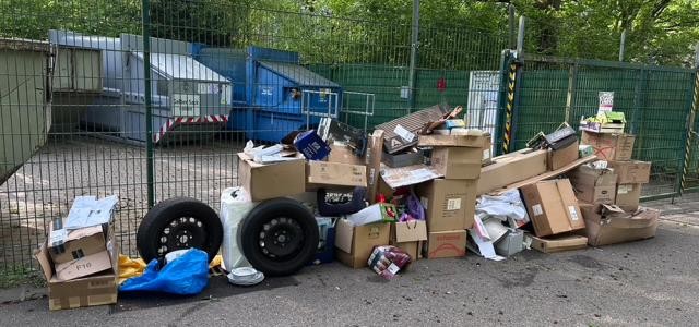 Kartons, Reifen und weiterer Unrat liegen vor einem Metallzaunzaun, dahinter Müllcontainer.