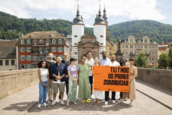 „Don’t let Europe go backwards”: Die Europäische Städtekoalition gegen Rassismus feiert vom 23. bis 25. September ihr 20. Jubiläum in Heidelberg und diskutiert lokale Strategien zum Schutz vor Diskriminierung.(Foto: Joe Pohl) Eine Gruppe von Menschen steht mit einem orangenen Plakat auf einer Brücke