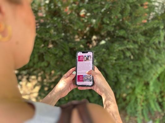 Die neuen Stadtteil-Rallyes in der MeinHeidelbergApp führen an Orte, die für junge Menschen interessant sind. (Foto: Stadt Heidelberg) Blick über die Schulter einer jungen Frau, sie bedient ein Smartphone mit geöffneter MeinHeidelbergApp, im Hintergrund ist verschwommen ein Busch zu sehen.