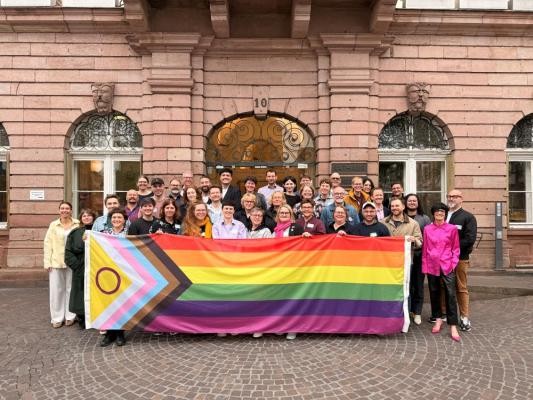 LSBTIQ+-Beauftragte aus ganz Deutschland tauschten sich beim Bundesnetzwerktreffen Queer.Kommunal vom 6. bis 8. Mai 2026 in Heidelberg über die Weiterentwicklung queerer Chancengleichheit in Bund, Ländern und Kommunen aus. (Foto: Stadt Heidelberg) LSBTIQ+-Beauftragte mit Regenbogen-Flagge vor dem Heidelberger Rathaus.
